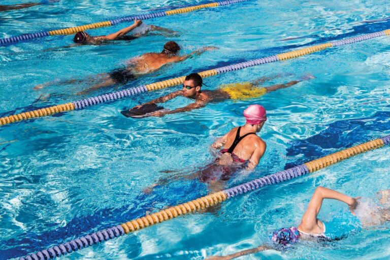 A group of men and women receiving swimming instruction together in a bright, indoor pool.